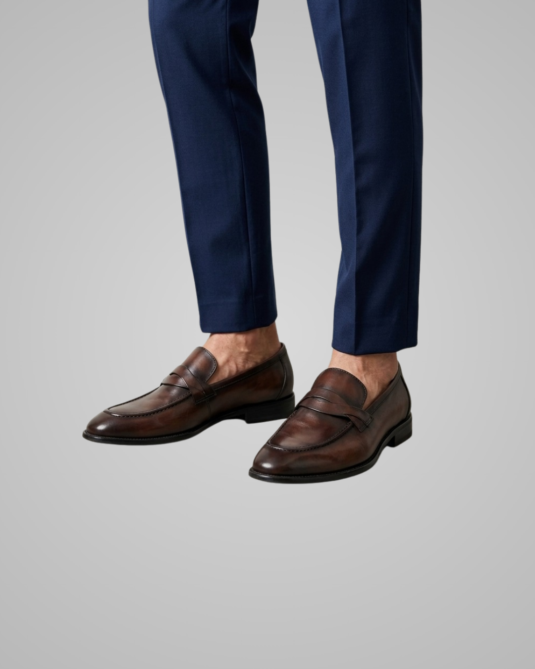 A close-up of a person's legs wearing dark brown leather penny loafers and navy blue trousers, shown against a solid light gray background.