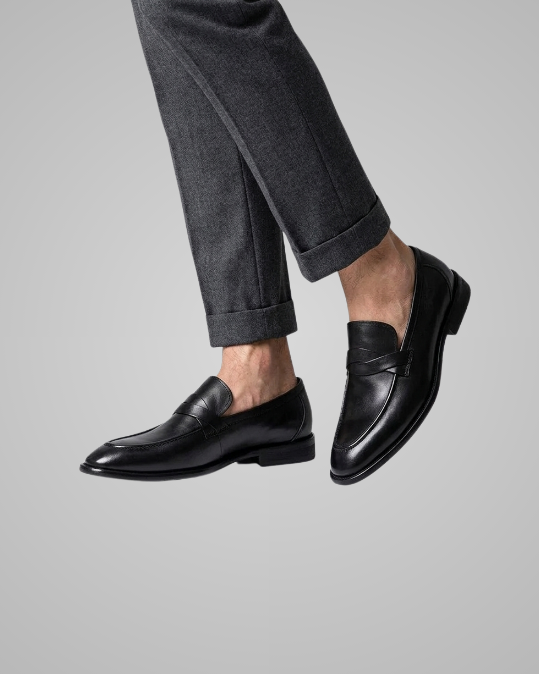 A pair of men's black leather penny loafers worn with dark gray cuffed trousers, shown against a solid light gray background.