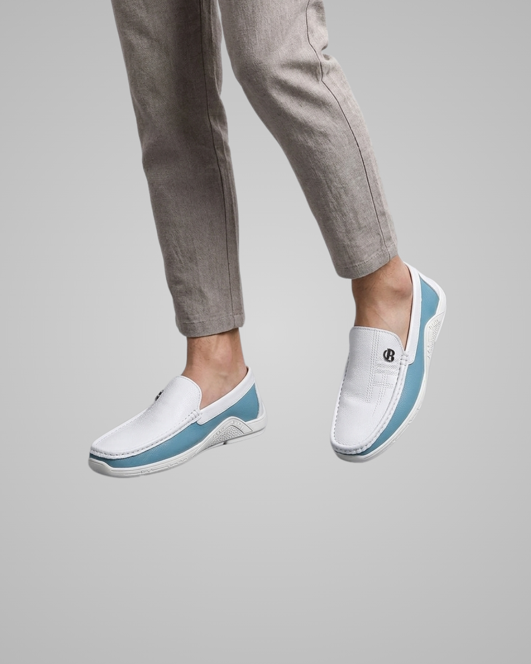 White slip-on loafers with a light blue side accent and a small black logo, shown on a model wearing light gray trousers against a solid gray background.