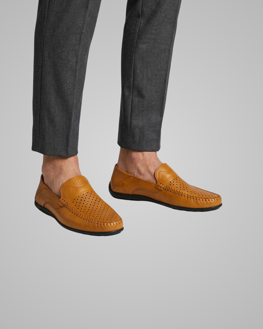 A pair of men's tan leather loafers with perforated detailing, shown on a model wearing dark gray trousers against a solid gray background.