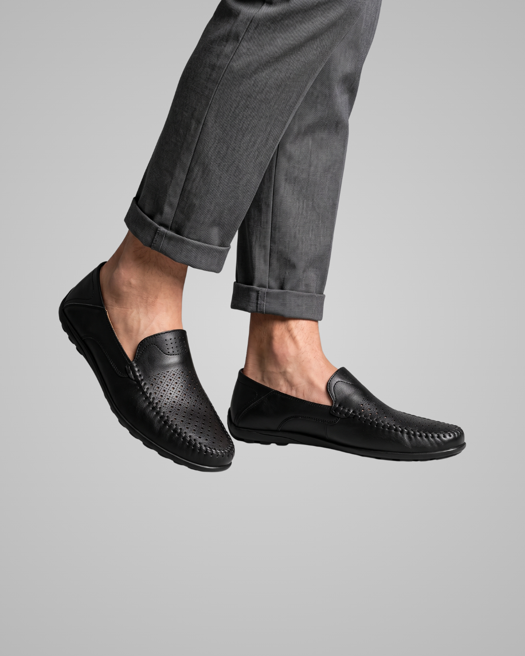 A pair of black leather slip-on loafers worn with cuffed gray trousers, shown against a solid light gray background.