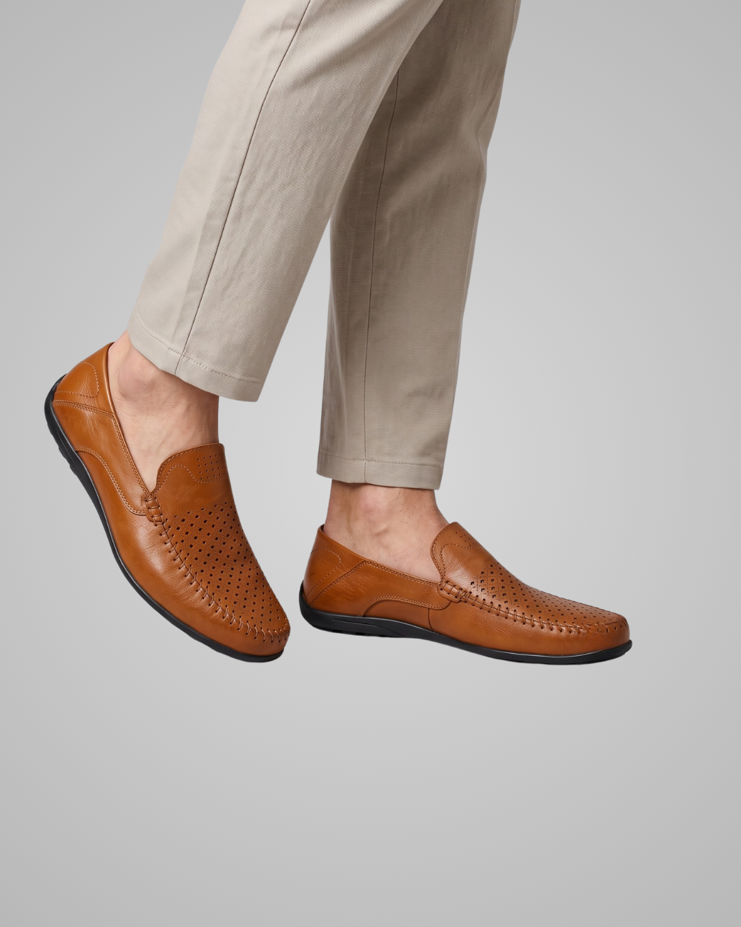A person wearing tan leather slip-on loafers with perforated detailing and light beige trousers, shown against a plain light gray studio background.