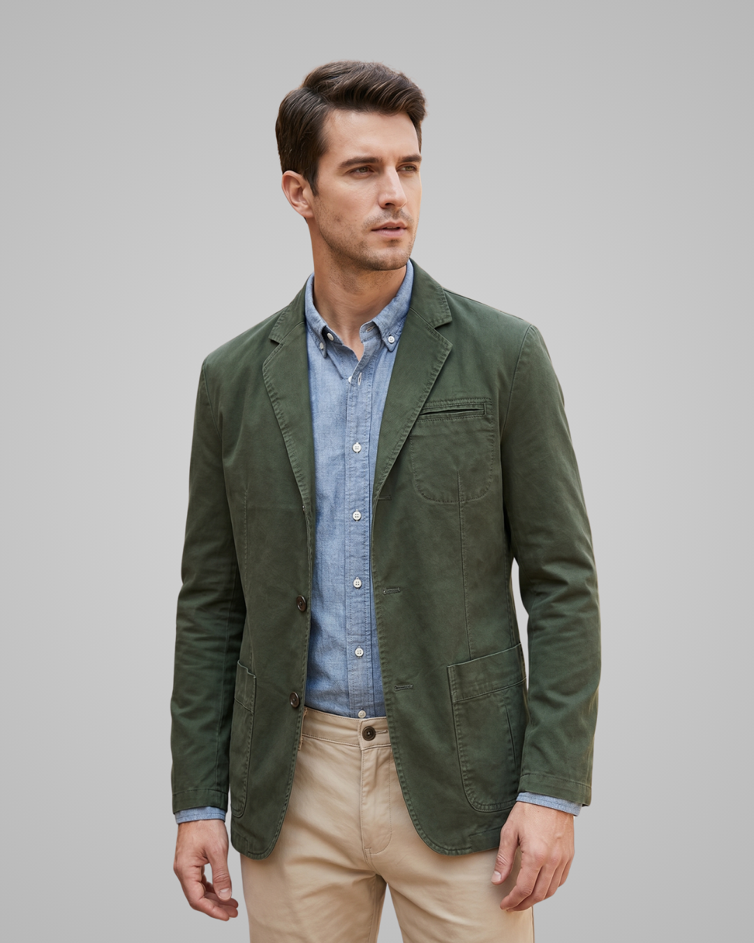 Man wearing a green jacket over a blue shirt and beige pants on a gray background