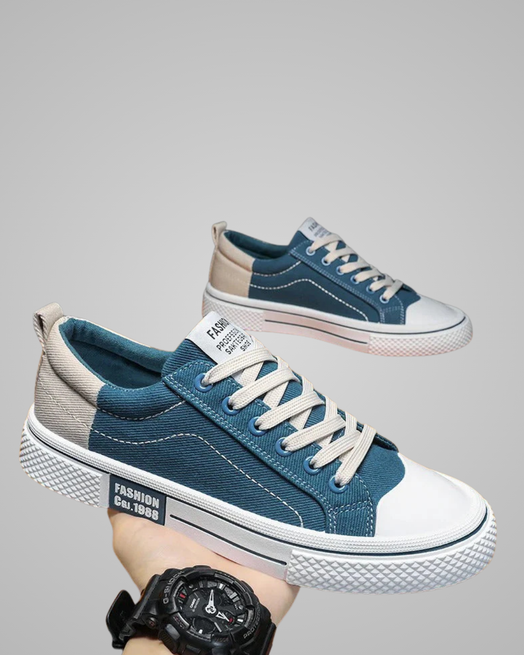 Blue and gray sneakers with white soles held by a hand wearing a black watch on a gray background