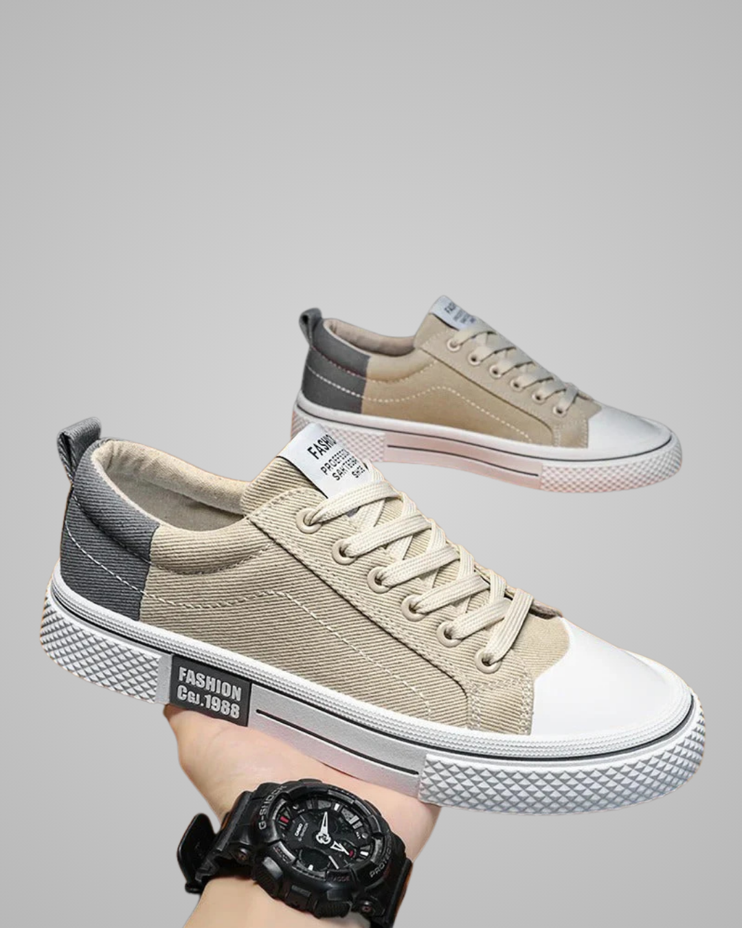 Pair of beige and gray sneakers with white soles held by a hand wearing a black watch.
