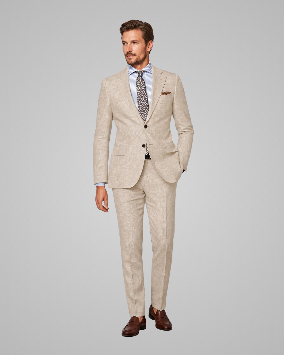 A man modeling a light beige two-piece suit with a subtle textured pattern, paired with a light blue dress shirt, a patterned tie, and brown leather dress shoes, set against a solid gray studio background.