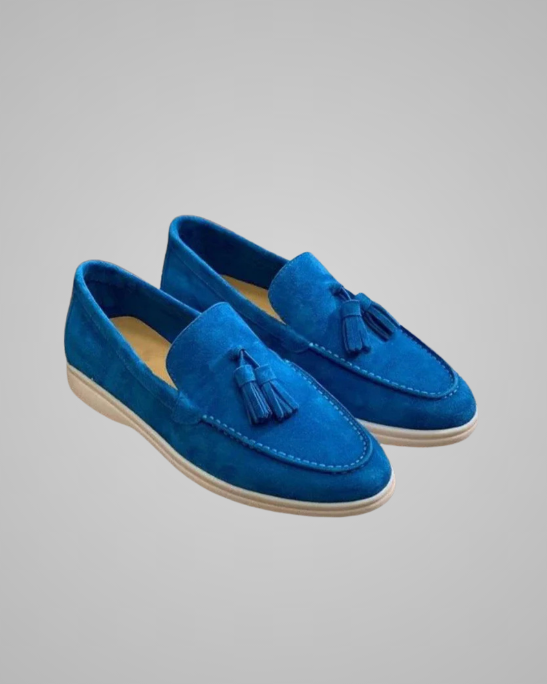 A pair of blue suede tassel loafers with off-white soles, shown on a neutral gray background.