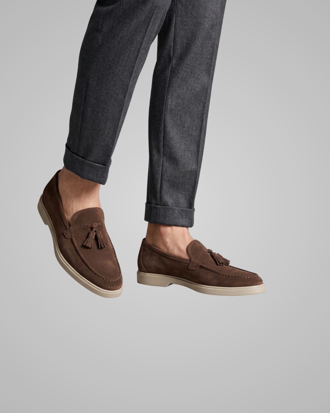 A person wearing dark brown suede tassel loafers with off-white soles, paired with dark gray cuffed jeans, shown against a solid light gray background.