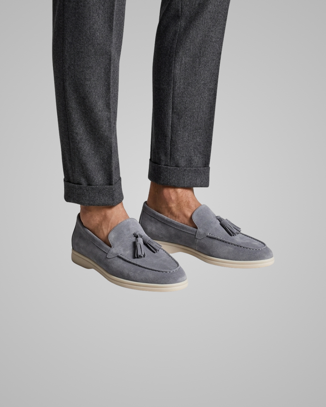 A person wearing gray suede tassel loafers with off-white soles and dark gray cuffed trousers, shown from the calves down against a light gray studio background.