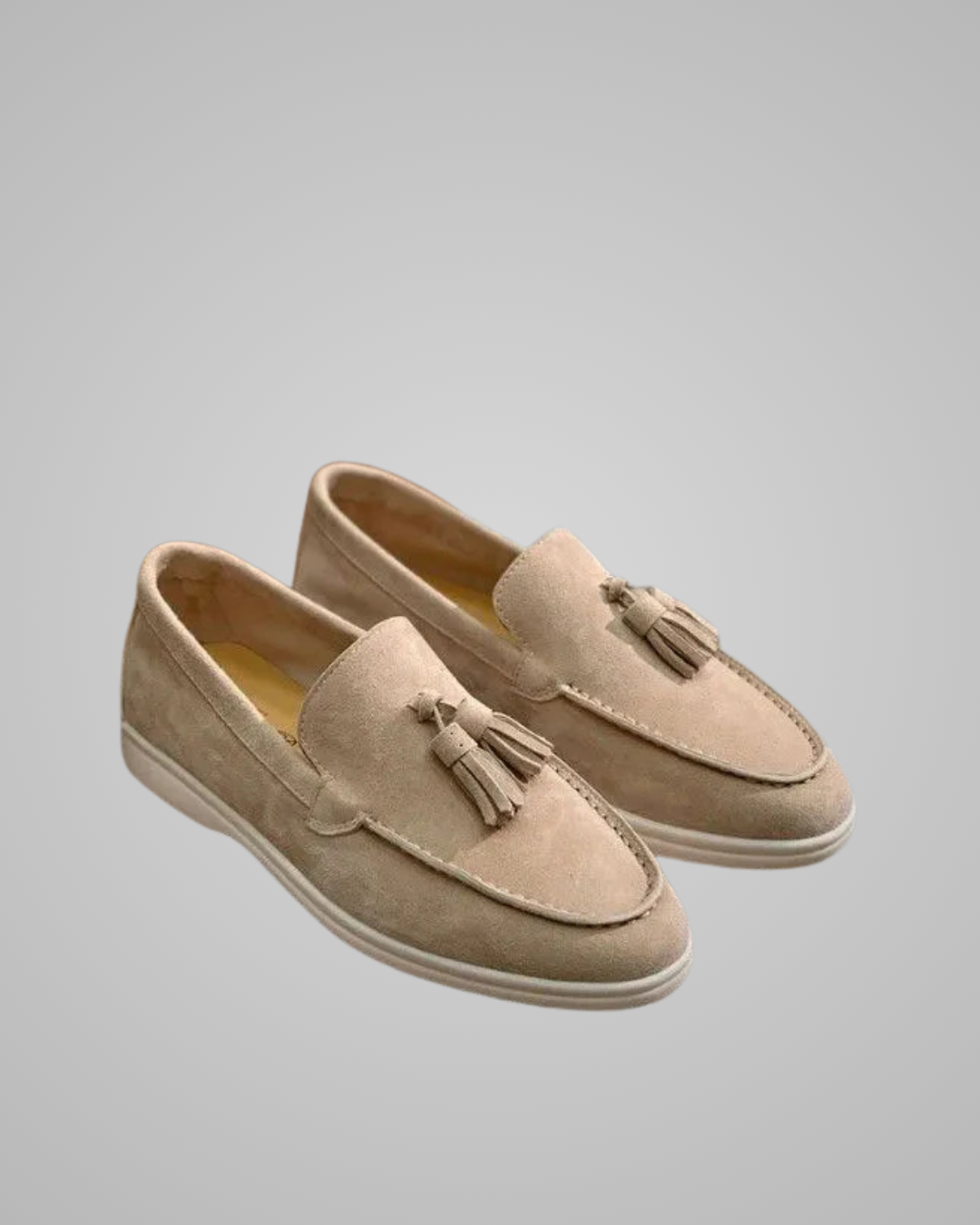A pair of beige suede tassel loafers with white soles, shown from a three-quarters angle on a neutral gray background.