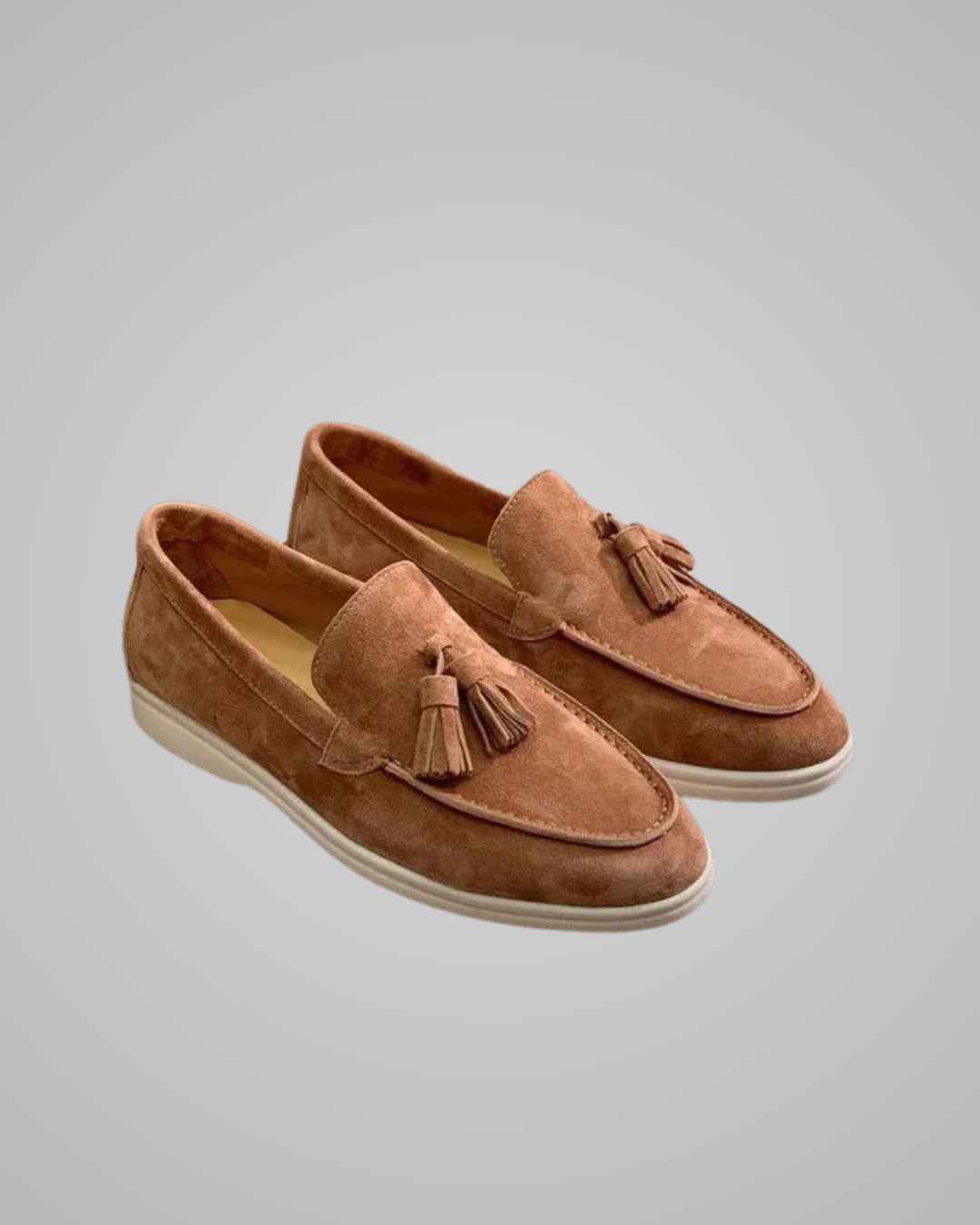 A pair of men's tan suede slip-on loafers with matching suede tassels and white soles, shown on a neutral gray background.