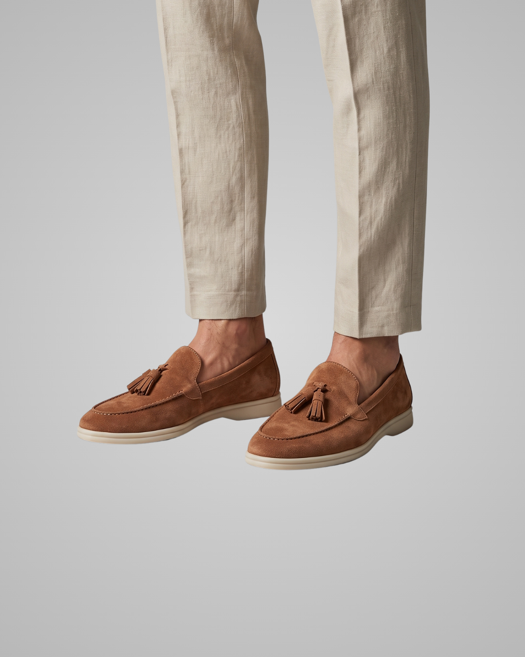 A pair of men's tan suede tassel loafers worn with light beige trousers, shown from the knees down against a solid gray background.