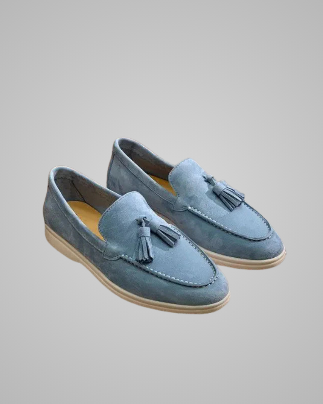 A pair of dusty blue suede tassel loafers with beige soles, displayed on a neutral gray background.