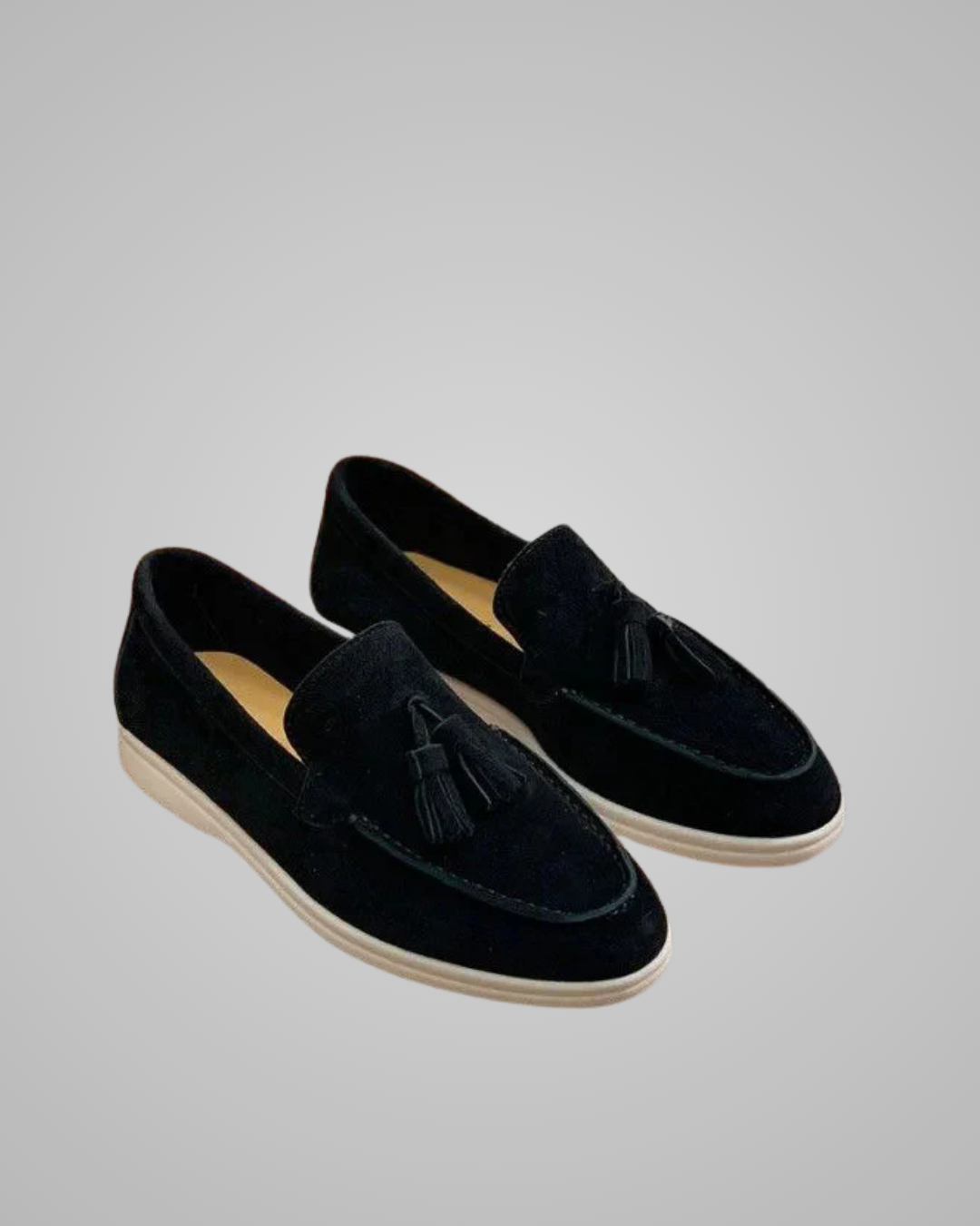 A pair of black suede tassel loafers with off-white soles, displayed on a neutral light gray background.