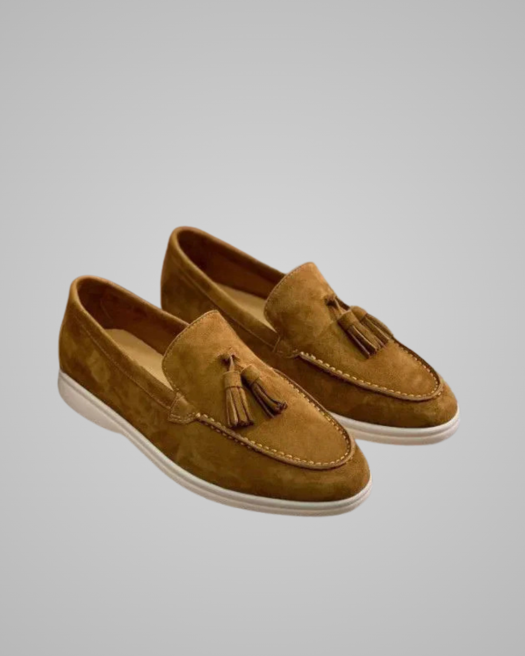 A pair of tan suede tassel loafers with white soles, displayed on a neutral light gray background.