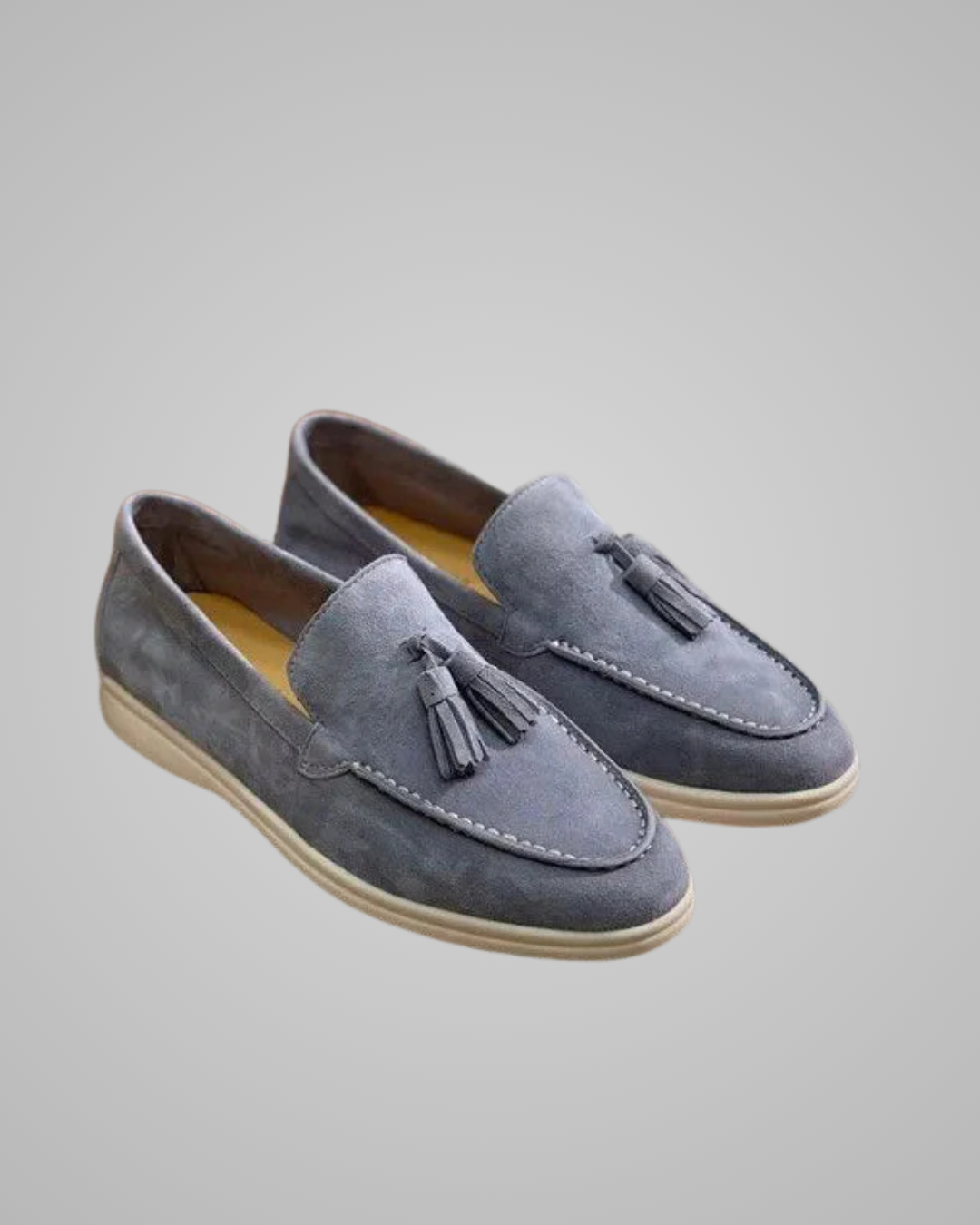 A pair of gray suede slip-on loafers with tassel details and light-colored soles, shown on a neutral gray background.