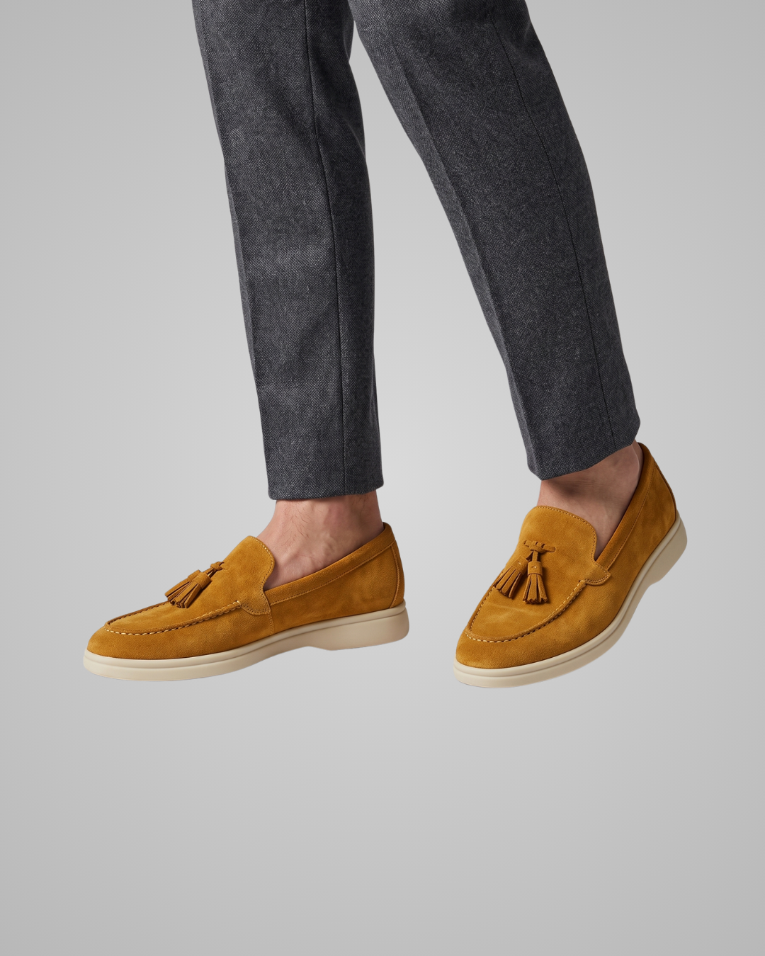 A pair of tan suede tassel loafers with white soles, displayed on a neutral light gray background.