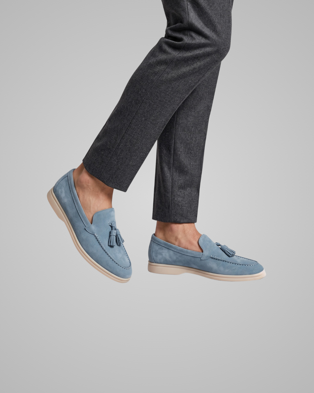 A person wearing light blue suede tassel loafers with dark gray trousers, shown from the knees down against a solid light gray background.