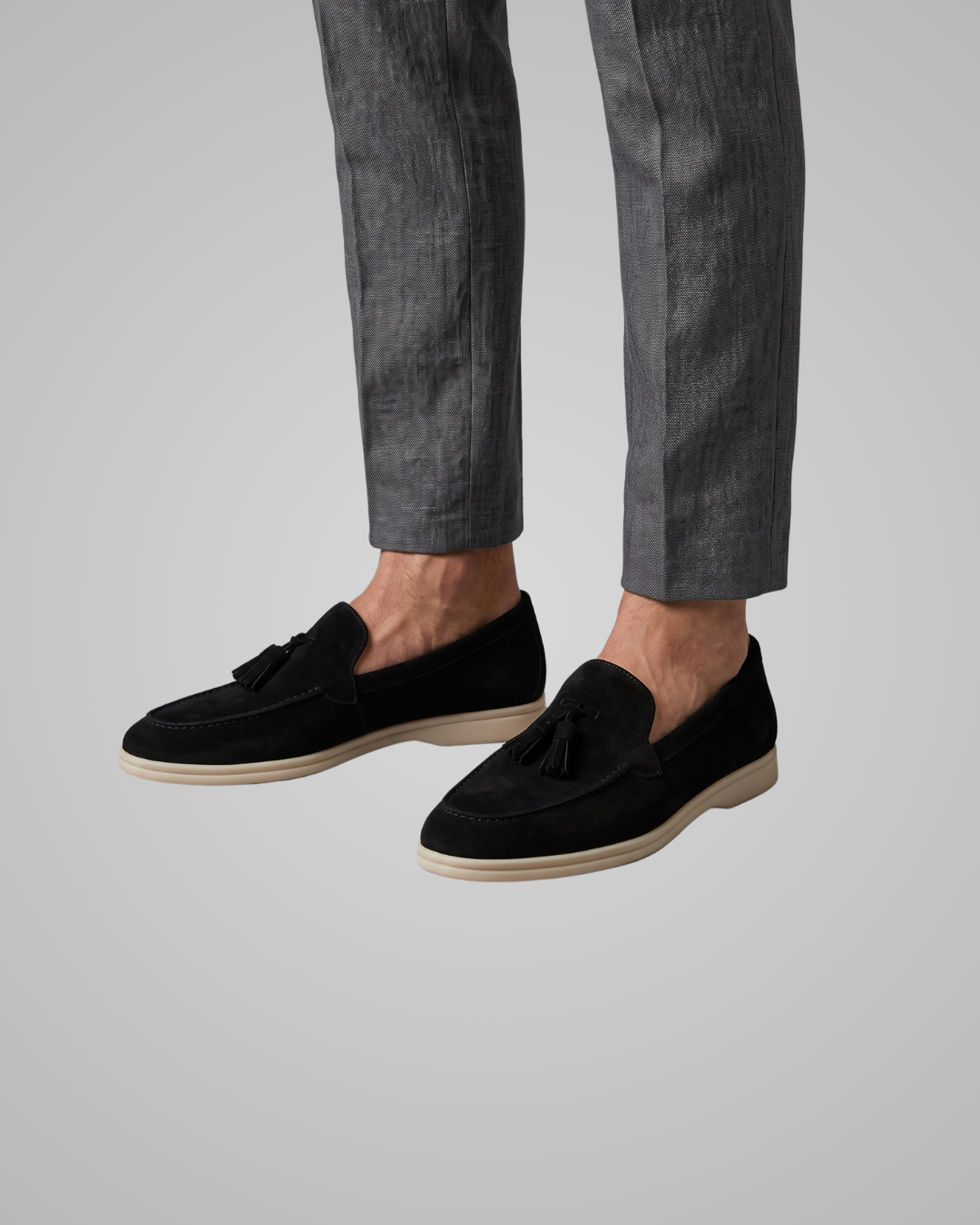 A pair of black suede tassel loafers with off-white soles, displayed on a neutral light gray background.