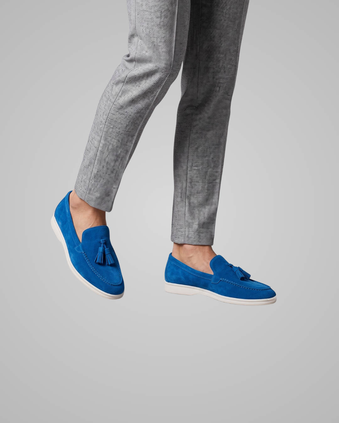A person wearing bright blue suede tassel loafers with white soles, paired with gray trousers, shown against a solid gray background.