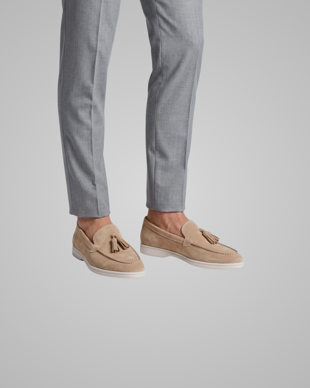 A person wearing light gray trousers and beige suede tassel loafers, shown from the knees down against a plain light gray studio background.