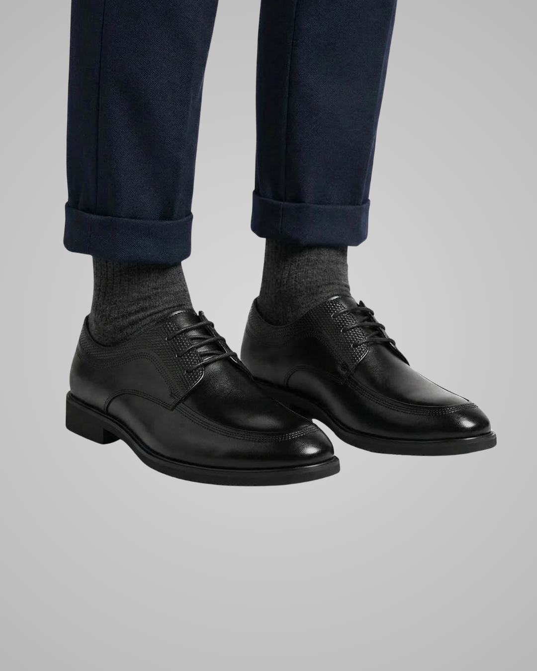 Black dress shoes worn with navy pants and gray socks on a light gray background