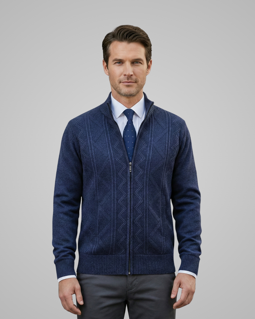 Man wearing a blue patterned sweater over a white shirt with a blue tie, standing against a gray background.