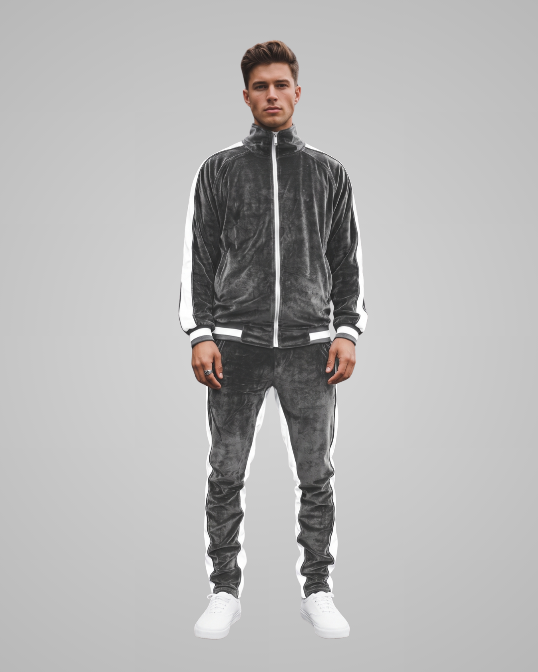 A man modeling a dark gray velour tracksuit with white side stripes, featuring a zip-up jacket and matching jogger pants, paired with white sneakers, against a solid gray background.