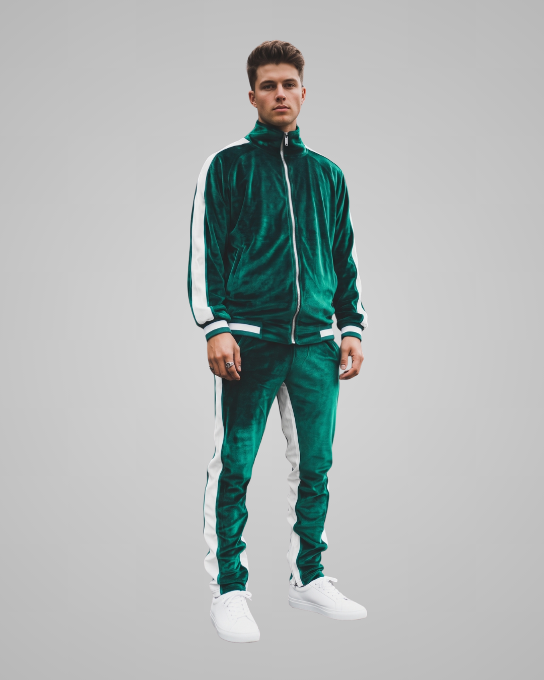A man modeling a dark green velvet tracksuit with white side stripes, featuring a zip-up jacket and matching pants, paired with white sneakers, against a solid gray background.