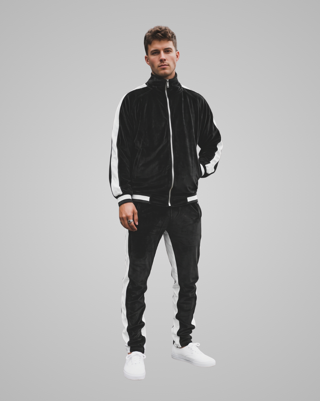 A man modeling a black velour tracksuit with white side stripes, featuring a zip-up jacket and matching jogger pants, paired with white sneakers, against a solid gray background.