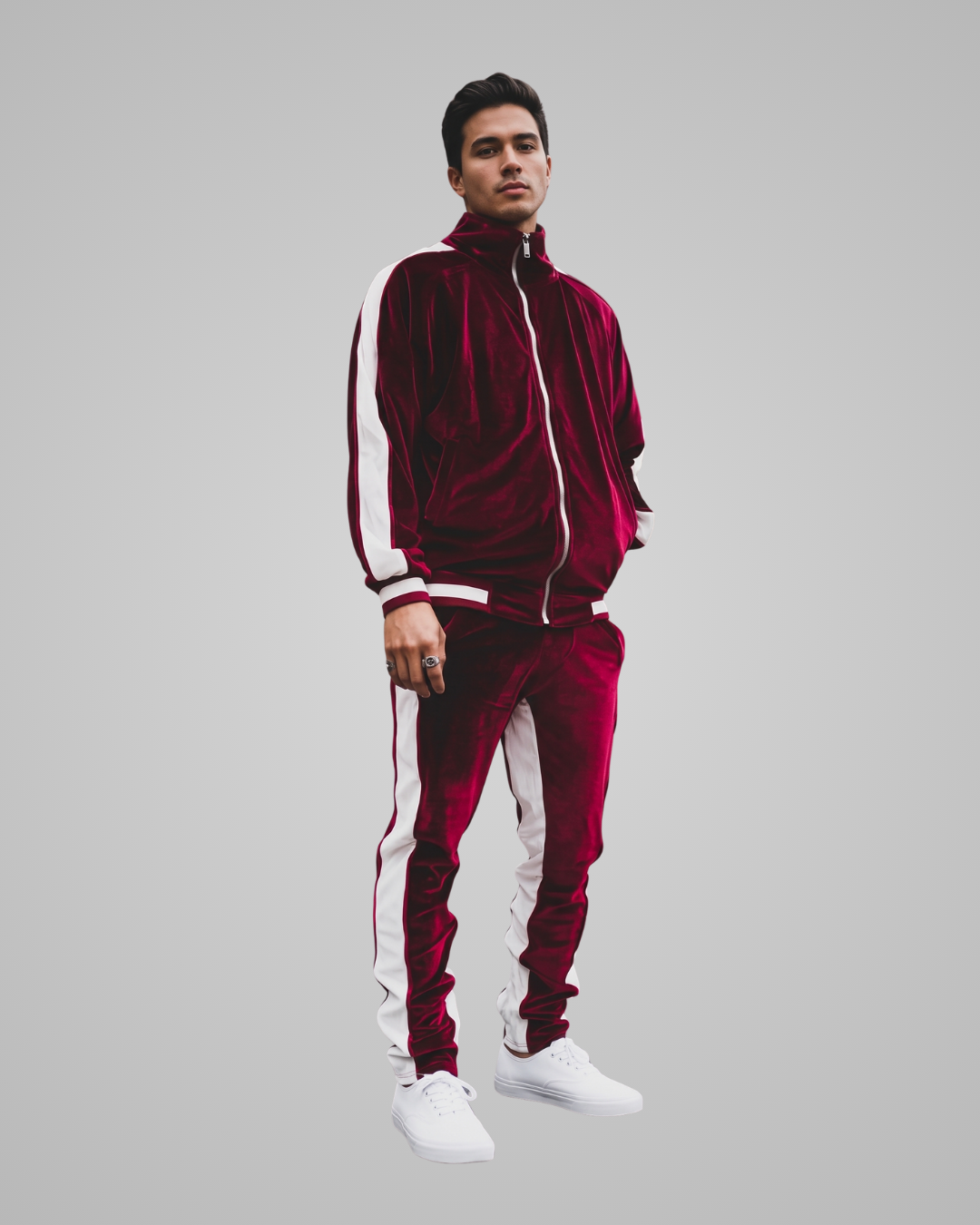 A man modeling a matching burgundy velvet tracksuit with white side stripes, featuring a zip-up jacket and matching pants, paired with white sneakers, against a solid gray background.