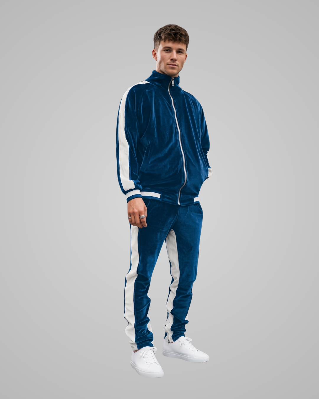 A man modeling a matching blue velour tracksuit with white side stripes, consisting of a zip-up jacket and matching pants, paired with white sneakers, against a solid gray background.