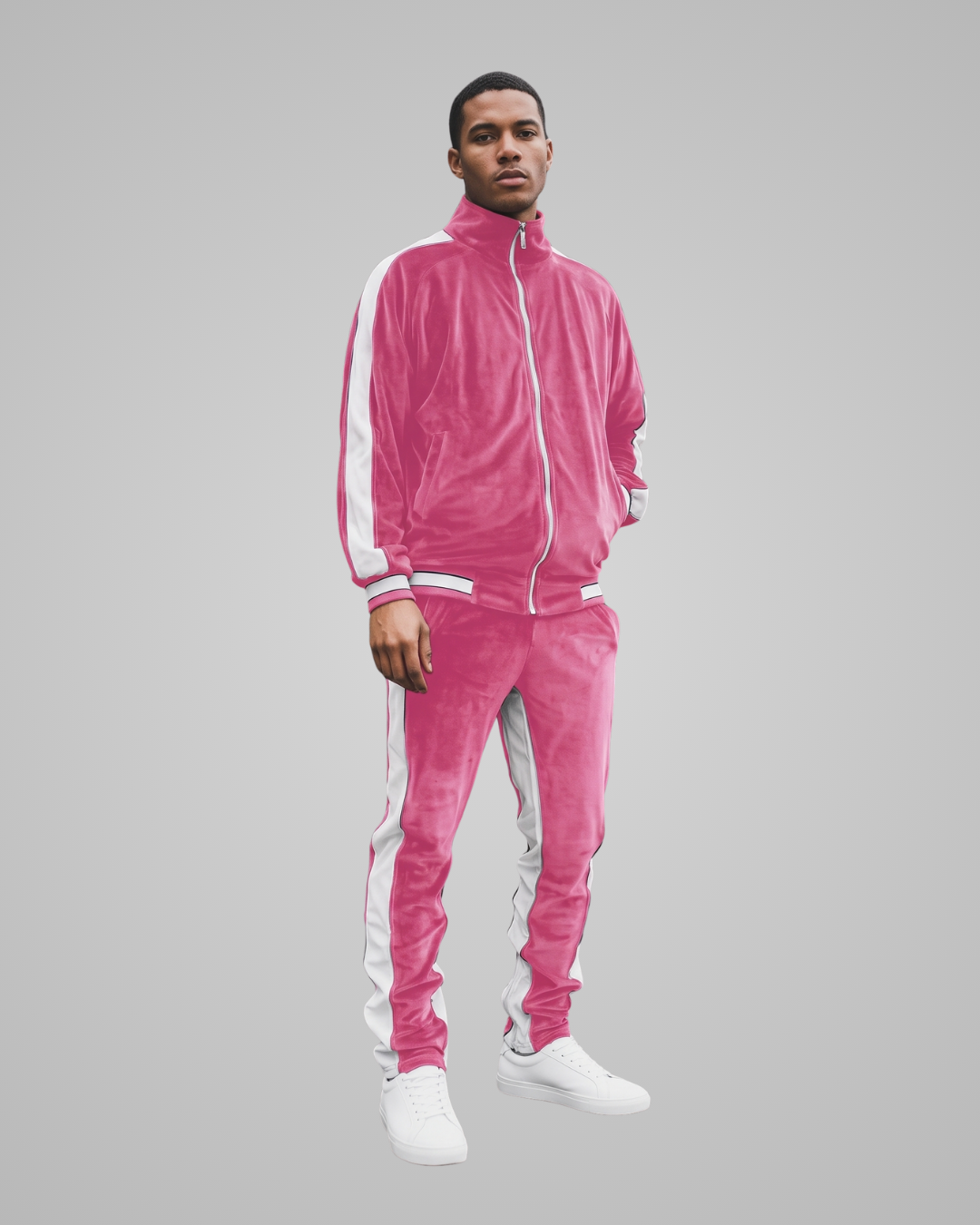 A man modeling a bright pink velour tracksuit with white side stripes and white sneakers, set against a solid gray studio background.