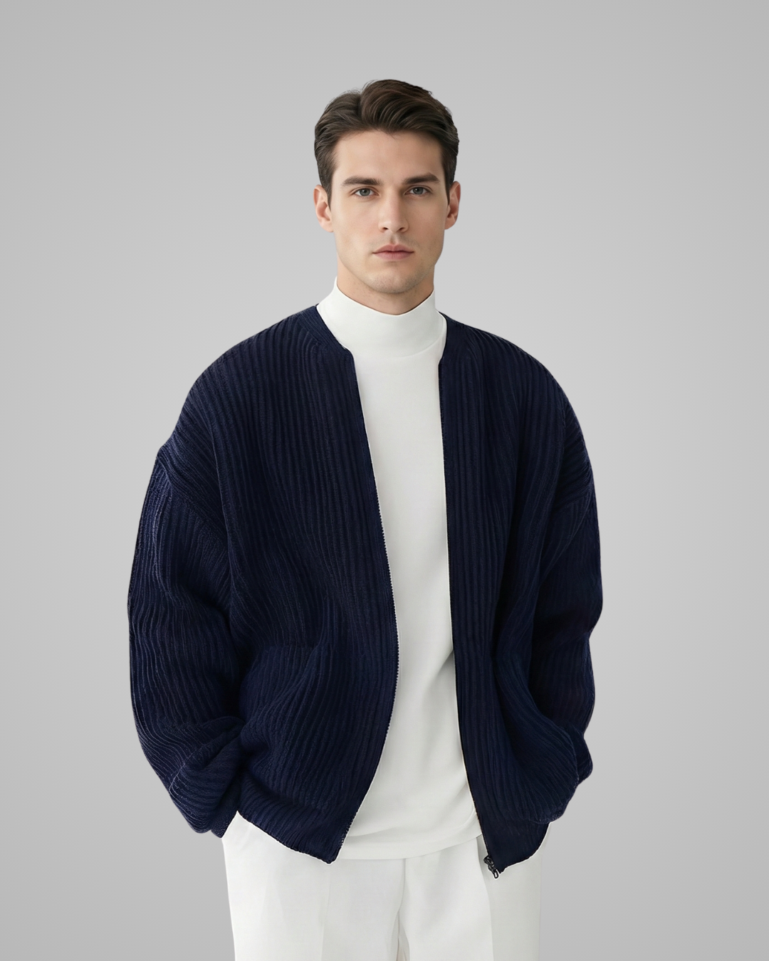 Man wearing a navy cardigan over a white turtleneck and pants on a gray background