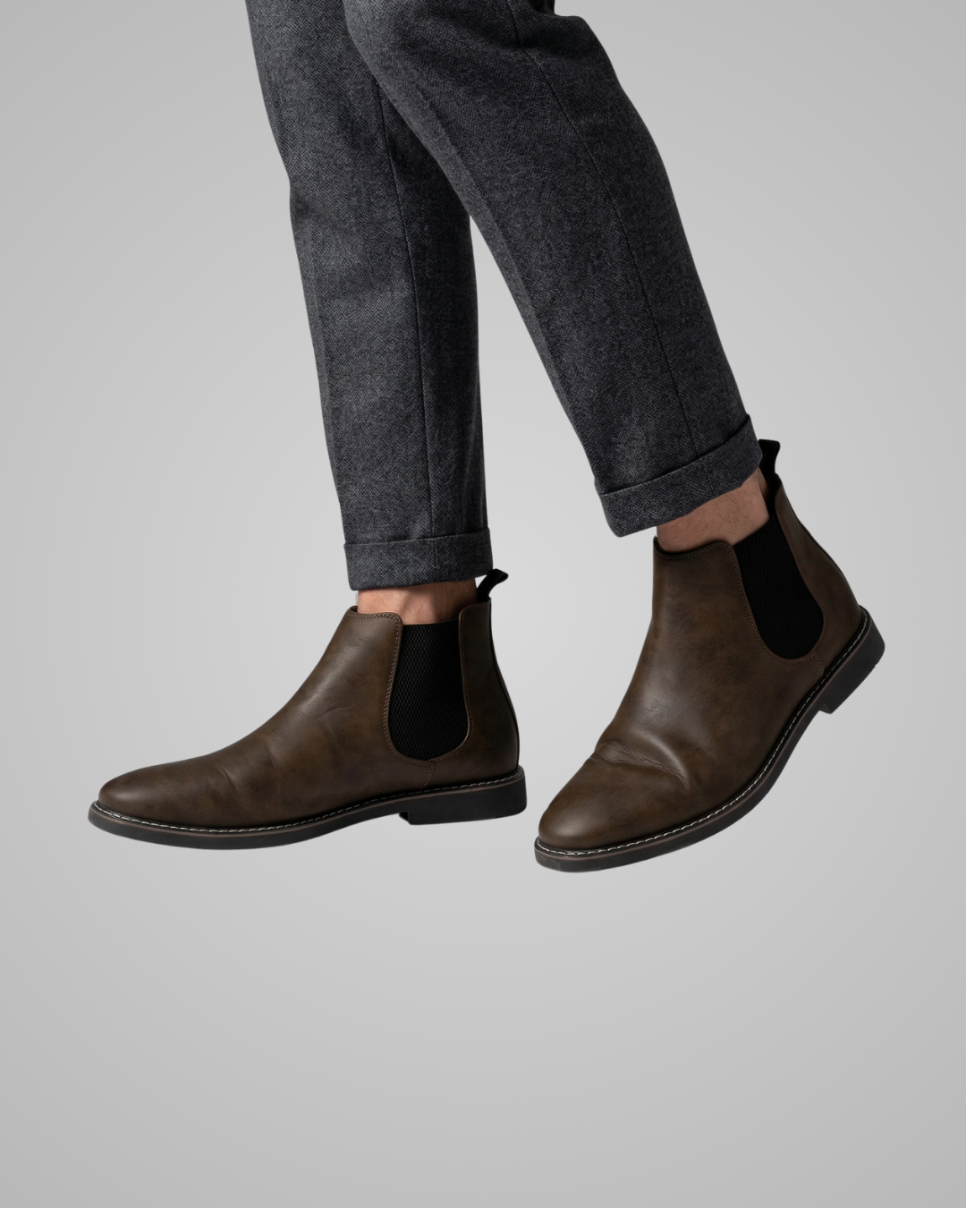 Brown leather Chelsea boots worn with rolled-up dark jeans on a gray background