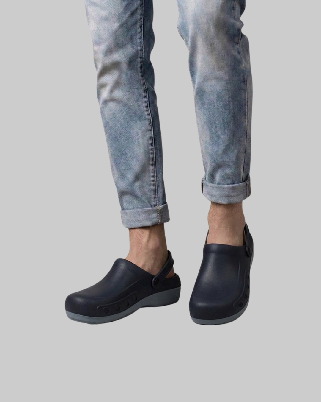 A person wearing light blue cuffed jeans and black slip-on clogs against a plain light gray background.