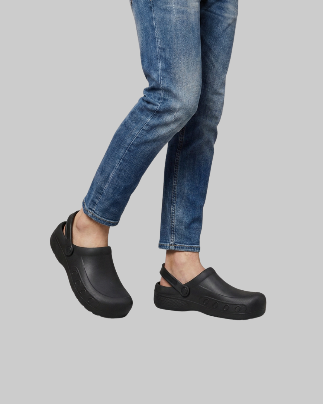 A person wearing blue jeans and black clogs against a solid gray background.