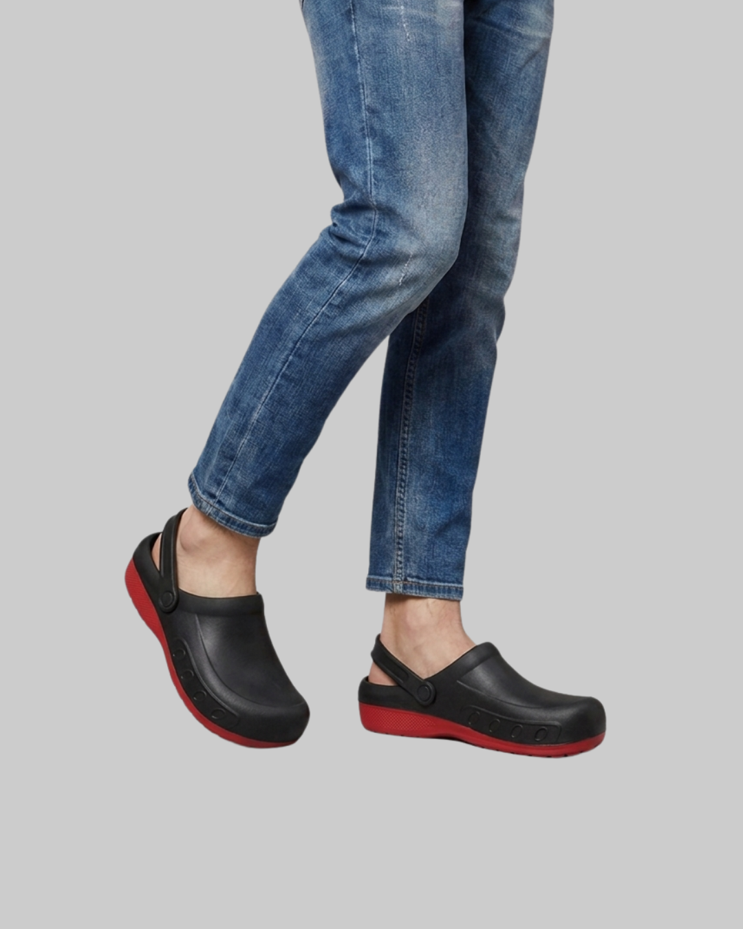Black clogs with red soles worn by a model in blue jeans, shown from the knees down against a solid gray background.
