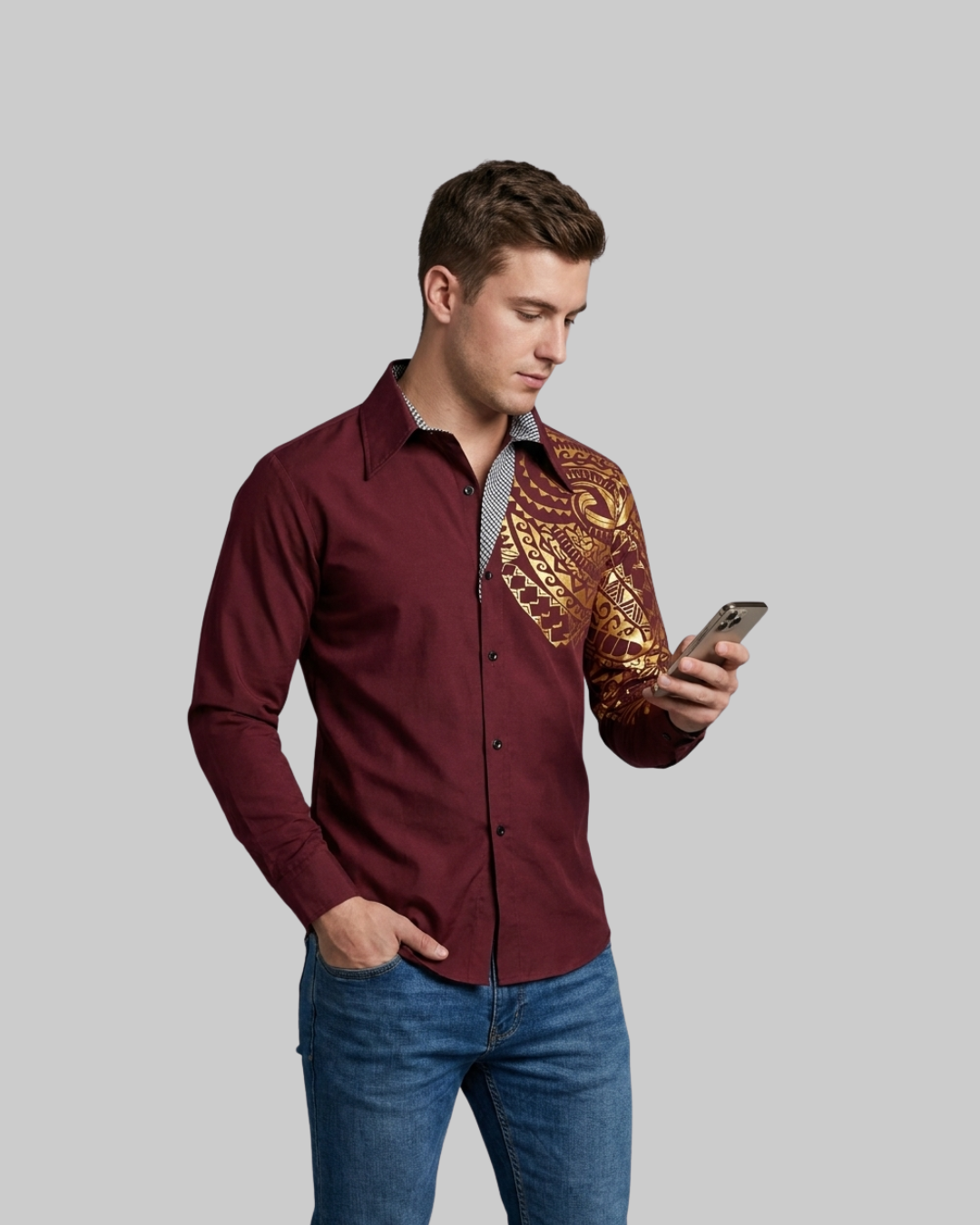 A man wearing a maroon long-sleeve button-up shirt with a gold and brown patterned sleeve, paired with blue jeans, holding a smartphone against a solid gray background.