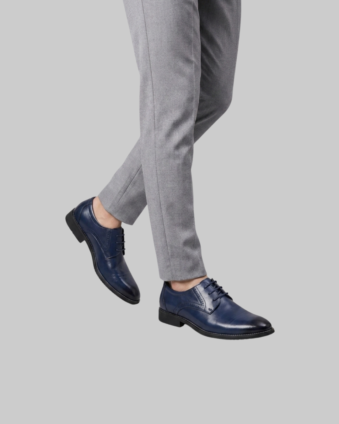 A person wearing navy blue leather oxford dress shoes with grey trousers, shown from the knees down against a solid light gray background.