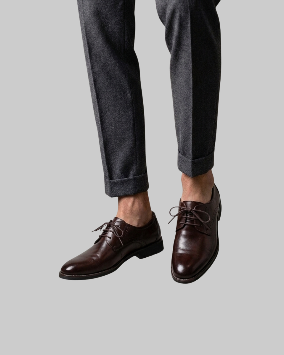 A pair of dark brown leather oxford dress shoes with laces, shown on a model wearing dark gray trousers against a solid gray background.