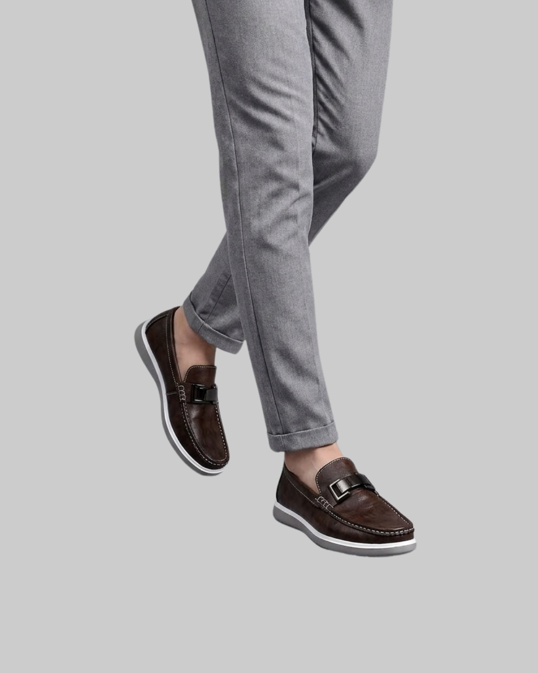 A person wearing gray pants and dark brown leather slip-on loafers with white soles, shown from the knees down against a plain light gray background.