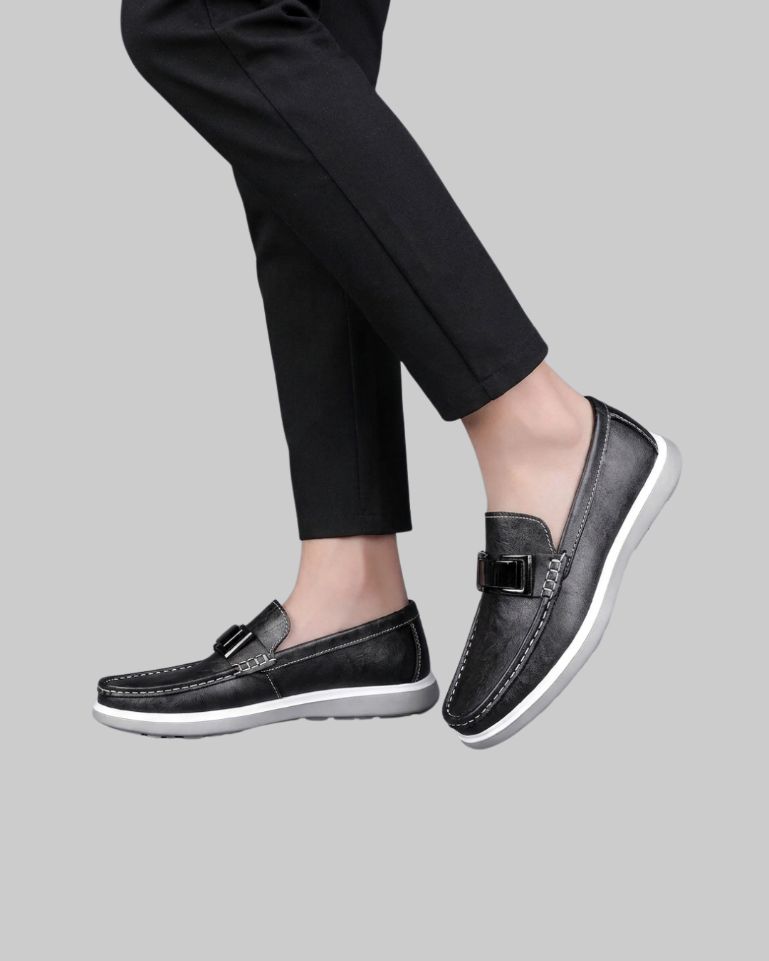 A pair of black leather slip-on loafers with a decorative buckle detail on the vamp and a white sole, shown on a model wearing black trousers against a solid gray background.