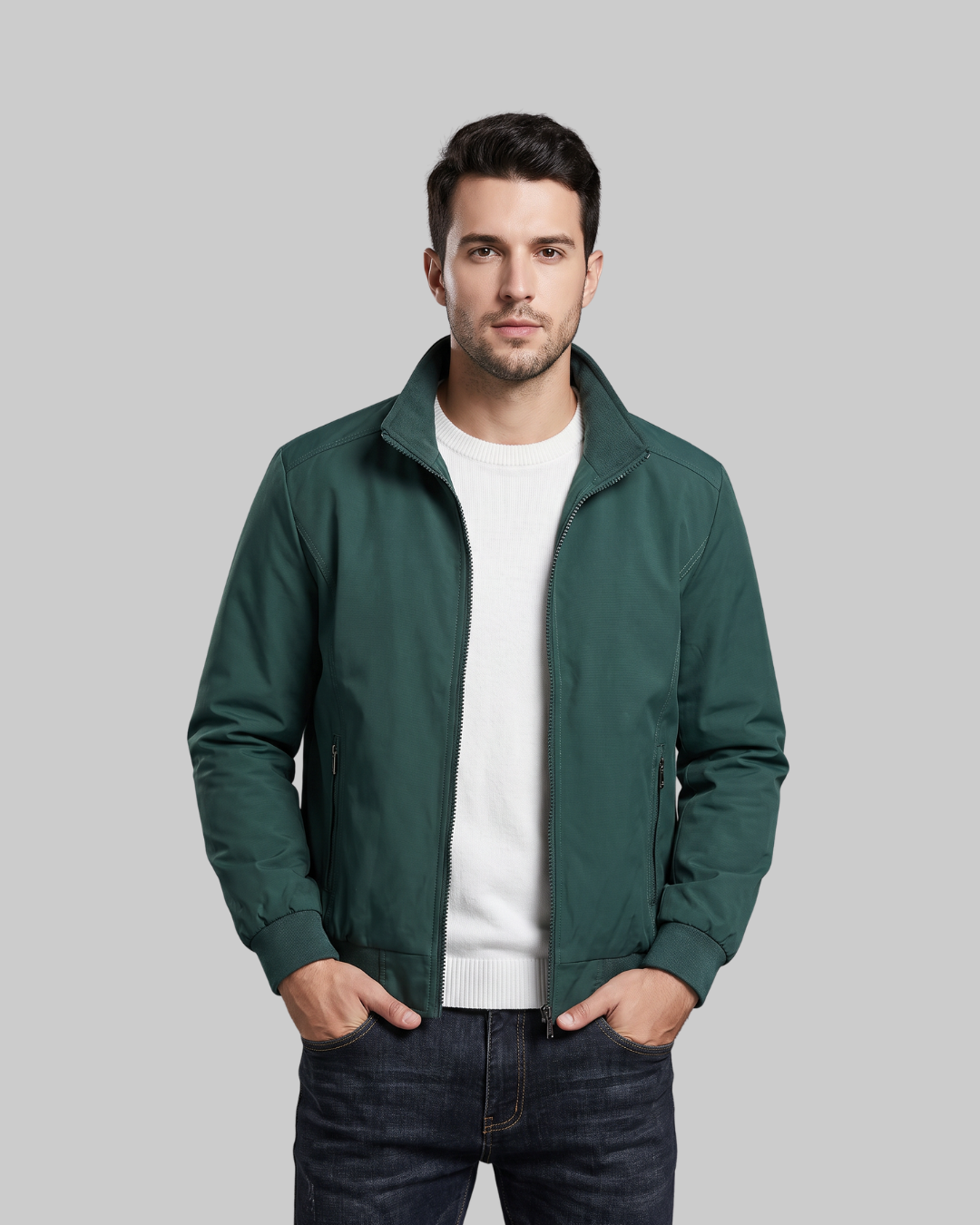 A man wearing a dark green zip-up jacket over a white crewneck sweater and dark jeans, standing against a solid gray background.