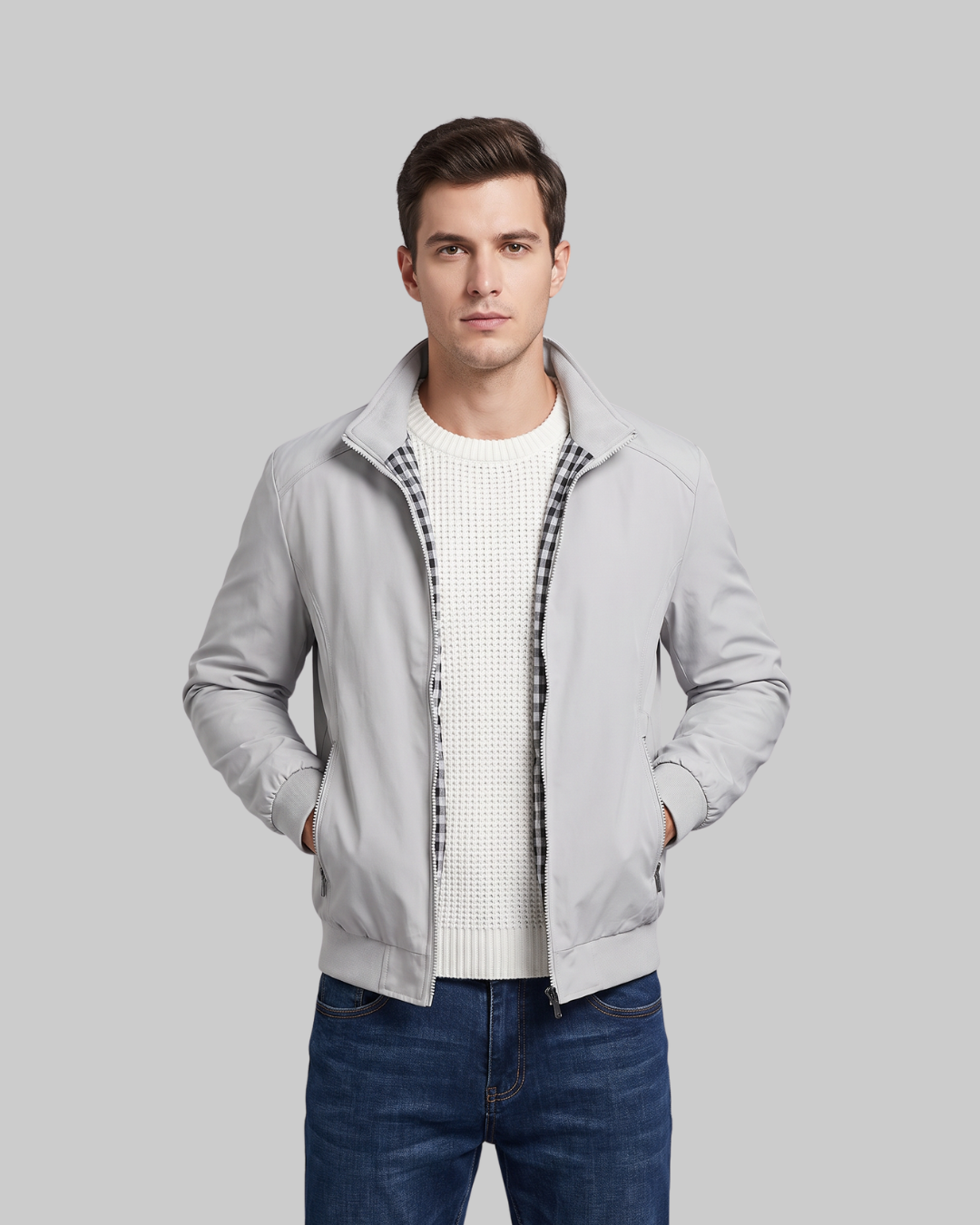 A man wearing a light gray zip-up jacket with a black-and-white checkered inner placket, layered over a white knit sweater and blue jeans, against a solid gray background.