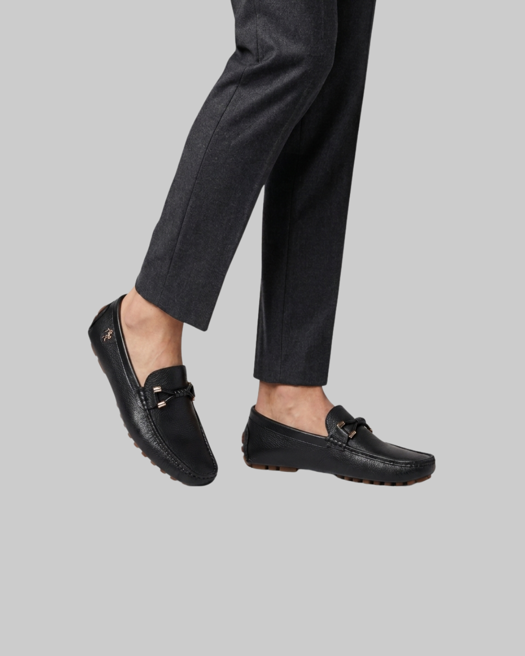Black leather penny loafers with a decorative strap and silver-tone buckle, shown on a model's feet against a solid gray background.