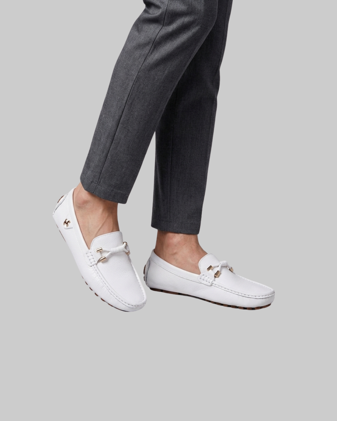 White leather driving loafers with gold-tone hardware and decorative ties, shown on a model wearing dark gray trousers against a solid gray background.