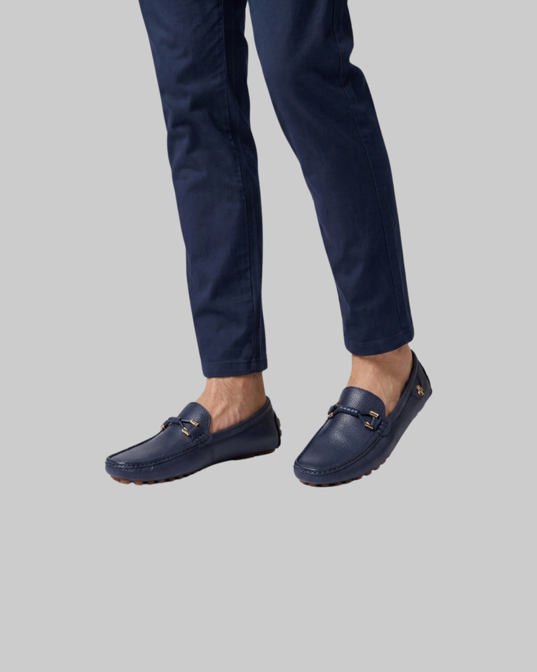 A person wearing navy blue leather loafers with gold-tone buckle details and matching navy trousers, shown from the calves down against a solid light gray background.