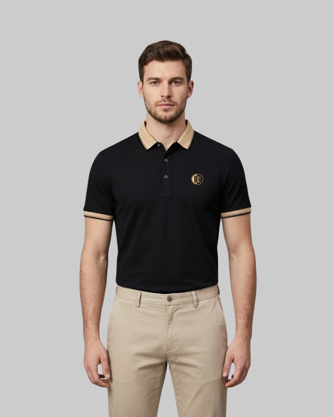 A man modeling a black short-sleeve polo shirt with beige collar and sleeve trim, paired with beige trousers, against a solid gray background.