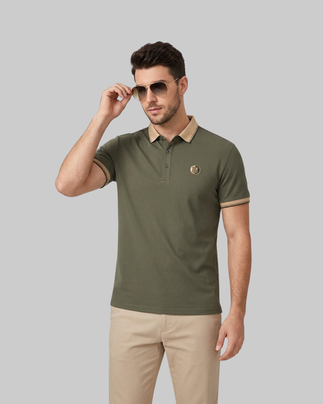 A man modeling an olive green short-sleeve polo shirt with beige trim on the collar and sleeve cuffs, paired with beige trousers, against a solid gray background.